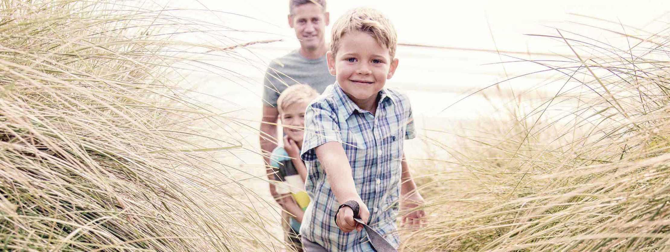 MS und ich: Ein Junge läuft mit seinem Hund durch die Dünen. Dahinten folgen sein kleiner Bruder und der Vater MS und ich: Ein Junge läuft mit seinem Hund durch die Dünen. Dahinten folgen sein kleiner Bruder und der Vater