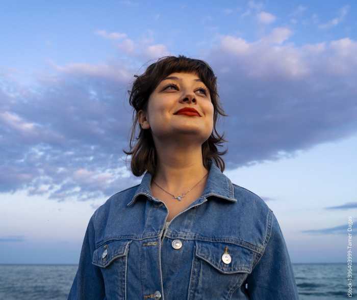 Eine junge Frau steht vor dem Meer und blick in den Himmel. 