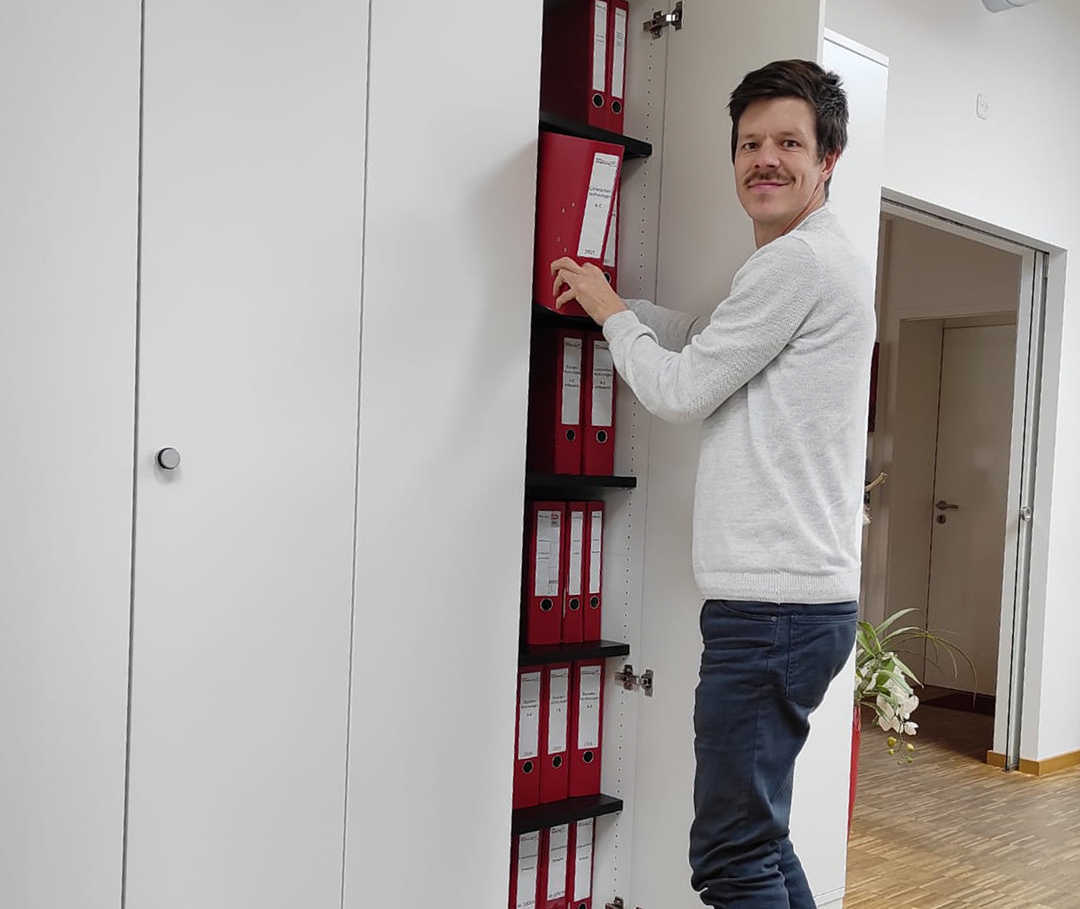 Das Bild zeigt Ricky stehend vor einem Aktenschrank
