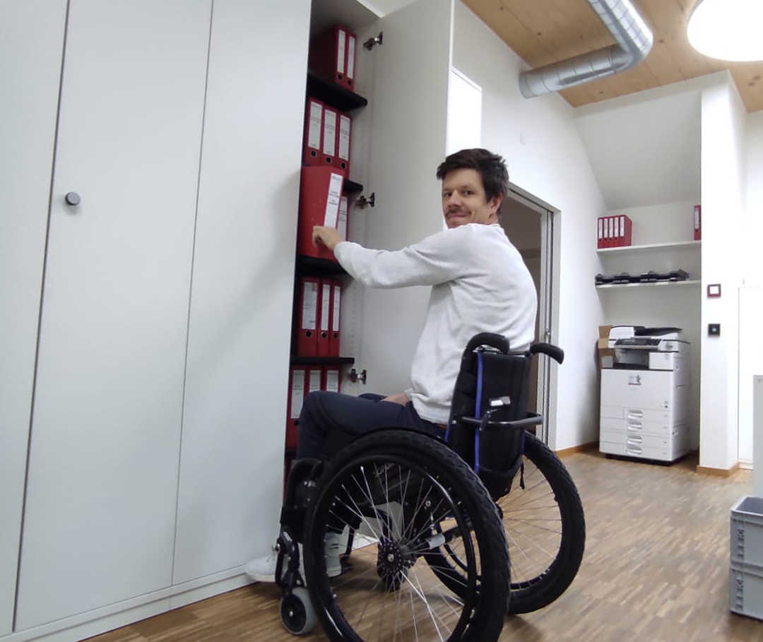 Das Bild zeigt Ricky im Rollstuhl vor einem Aktenschrank