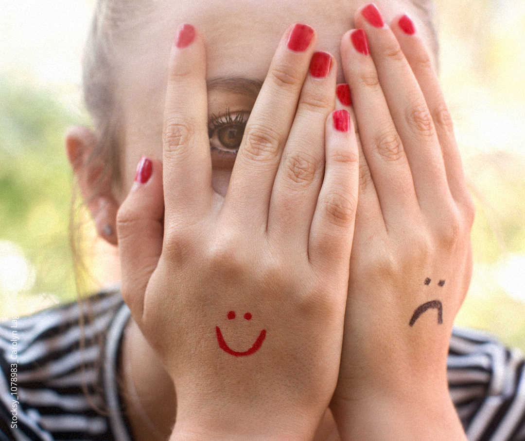 Zwei Hände vor einem Gesicht. Auf die eine Hand ist ein lächelndes Gesicht gemalt, auf die andere ein trauriges.
