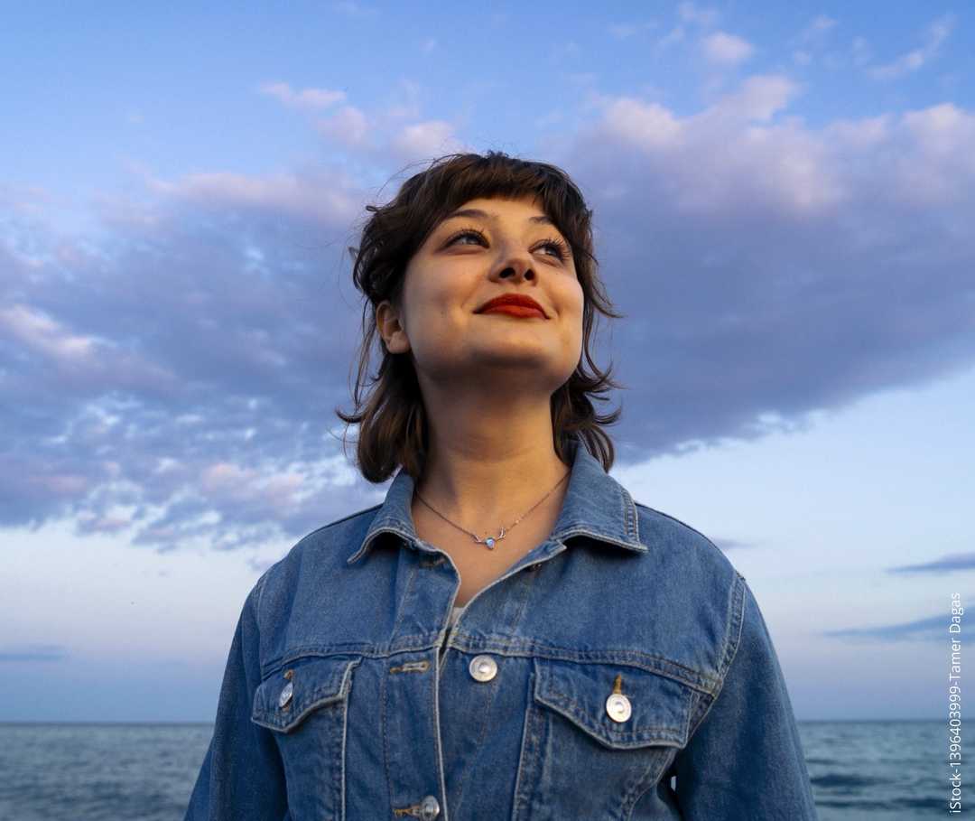 Eine junge Frau steht vor dem Meer und blick in den Himmel. 