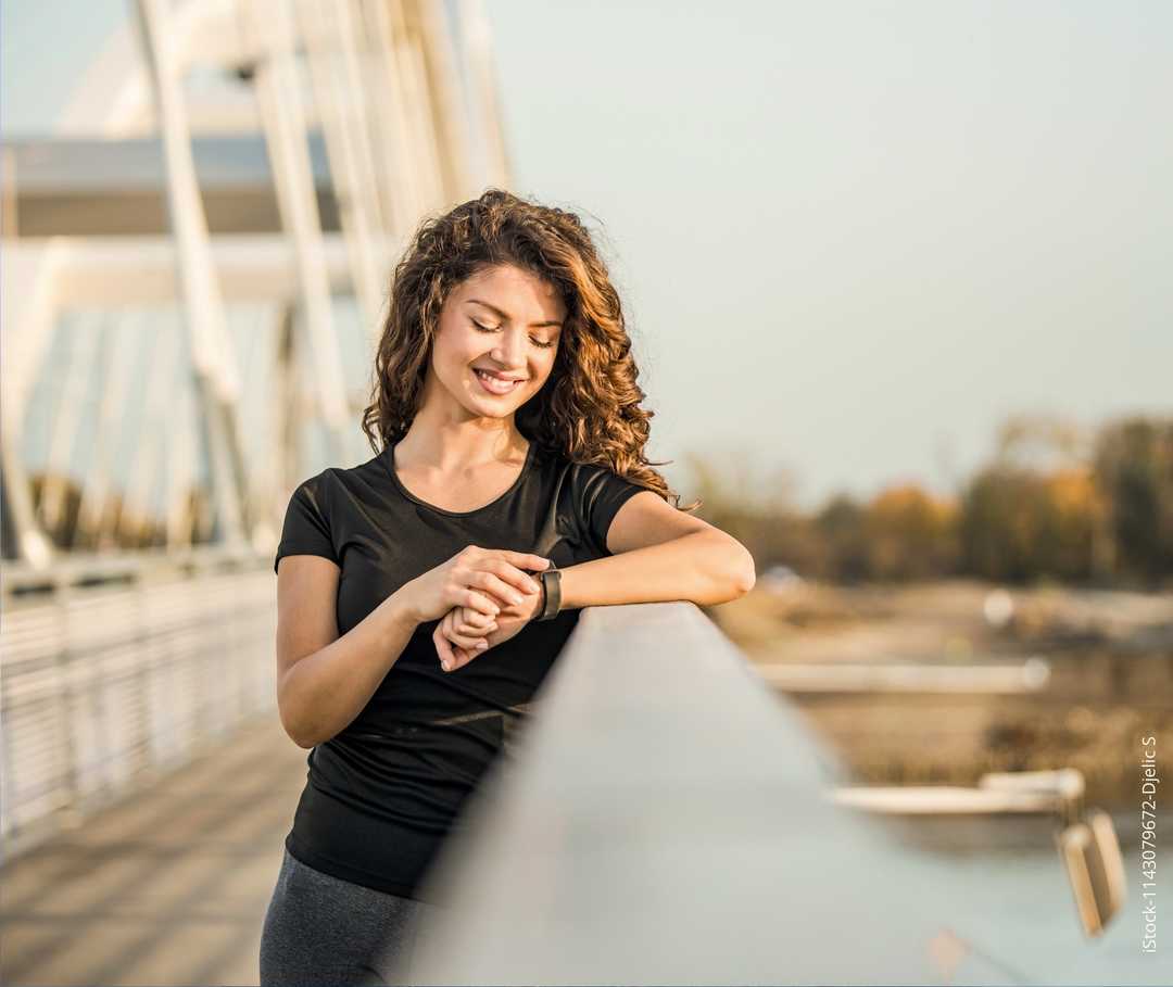 Eine junge Frau steht auf einer Brücke und schaut auf ihre Armbanduhr.