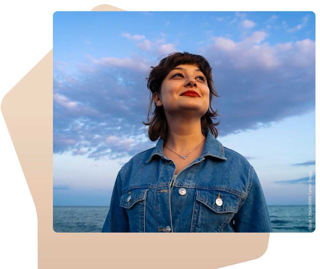 Eine junge Frau steht vor dem Meer und blick in den Himmel. 