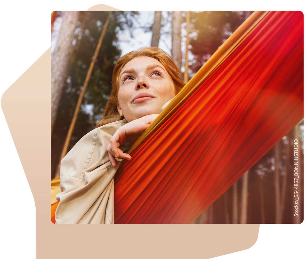 Eine Frau schaut verträumt in den Himmel während sie in einer Hängematte im Wald liegt.